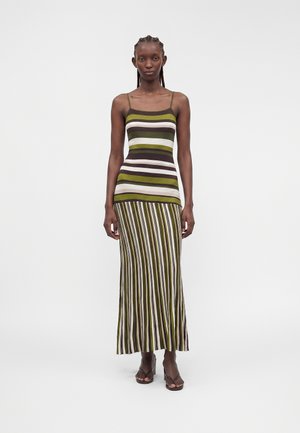 Mujer de pie, vistiendo un vestido sin mangas con rayas horizontales en la parte superior y rayas verticales en la falda en tonos verdes, blancos y marrones.