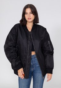 Młoda kobieta ubrana w czarną, oversize'ową kurtkę bombowiec, czarny top na krótki rękaw i niebieskie dżinsy, stojąca na jednolitym, jasnoszarym tle.