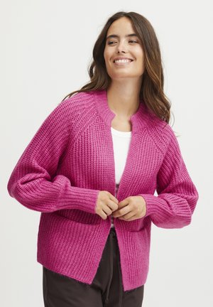 Cardigan tricoté en rose vif avec un motif texturé, à l'avant ouvert et avec de longues manches côtelées, porté sur un top blanc.