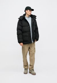 Fekete puffdzseki kapucnival, egy szürke kötött pulóver fölé rétegezve, bézs cargo nadrággal és szürke csizmával párosítva.