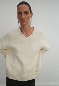 Maglione cream a vestibilità ampia con scollatura a V, polsini e orlo a coste. Texture morbida, maglia leggera, indossato sopra pantaloni scuri. Design minimalista.