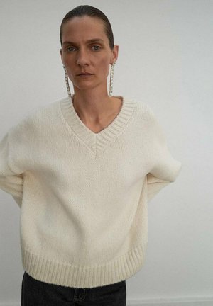 Suéter crema de corte holgado con escote en V, puños y dobladillo de canalé. Textura suave, tejido ligero, llevado sobre pantalones oscuros. Diseño minimalista.