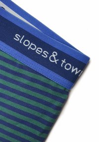 Tkanina w paski w odcieniach zieleni i granatu, z teksturowanym niebieskim pasem w talii oznaczonym napisem "stoki i ręczniki". Zbliżenie na tkaninę.