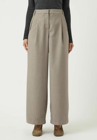 Brede pantalon van lichtgrijze stof, met voorplooien, een hoge taille en zijzakken. Gecombineerd met lage bruine schoenen.