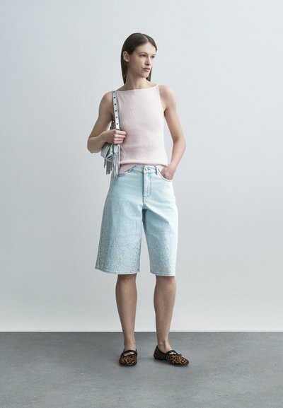 Γυναίκα με ίσια καστανά μαλλιά φοράει αμάνικο τοπ απαλό ροζ, ανοιχτόχρωμα τζιν σορτς μέχρι το γόνατο, παπούτσια με λεοπάρ σχέδιο και κρατά μικρή ασημένια τσάντα με φούντες.