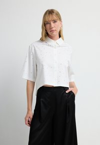 Camisa blanca corta con mangas cortas, cierre de botones y acentos plateados dispersos. Combinada con pantalones de pierna ancha negros.