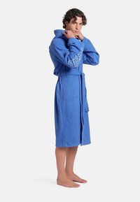 Blå frottébadrock med huva, midjebälte och två framfickor. Har broderad logotyp på ärmen. Mjuk textur och lätt i vikt.