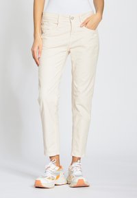 Pantalones de algodón beige con un corte ajustado, que cuentan con bolsillos frontales y cierre de botón. Combinados con zapatillas deportivas blancas y naranjas.