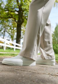 Weiße Slip-On Sneaker mit einer glatten, matten Oberfläche und subtilen Nahtdetails, kombiniert mit hellbeigen, leicht taillierten Hosen.