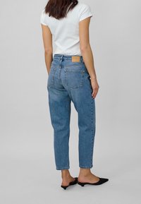 Höga blå denimjeans med avslappnad passform, fem fickor och en tan läderpatch på baksidan av midjebandet. Slät, något blekt textur.