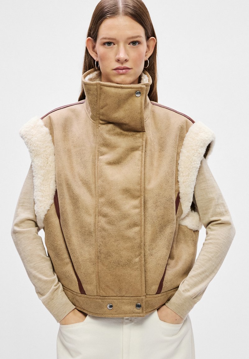 Beige Wildlederweste mit hohem Kragen, Lammfellakzenten an den Schultern und kastanienbraunen Seitenpanelen. Mit Druckknopfverschluss am unteren Ende.