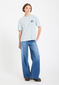 Jeune femme debout portant un t-shirt graphique bleu clair, un jean bleu à jambes larges et des chaussures marron à semelles épaisses, sur fond blanc.