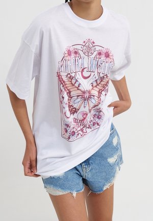 Persoon die een wit T-shirt draagt met een grote roze vlinder, bloemen en de tekst "Aurora Mystic", gecombineerd met gescheurde denimshorts.