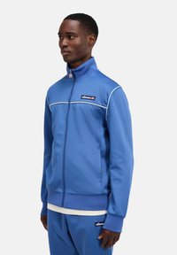 Blaue Sportjacke mit hohem Kragen, Reißverschluss und weißen Akzentstreifen. Mit Logopatch auf der Brust und gerippten Bündchen.