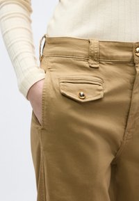 Άτομο που φοράει μπεζ παντελόνια με κουμπωτό καπάκι τσέπης μπροστά, με το χέρι μέσα στην πλαϊνή τσέπη, και ένα ελαφρύ μπεζ ριγωτό μακρυμάνικο τοπ.