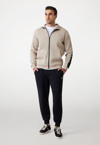 Giacca beige con zip e strisce laterali nere, abbinata a pantaloni jogger neri con logo e orli affusolati, completata da sneakers nere.
