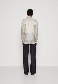 Blusa de seda en color crema con un reverso texturizado y un cuello decorativo, combinada con pantalones de encaje azul marino con un diseño de pierna acampanada.