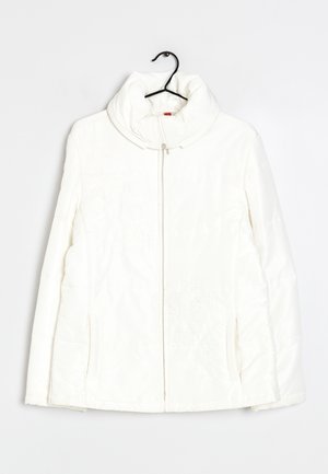 Veste matelassée blanche avec col montant et fermeture éclair frontale accrochée sur un cintre noir sur un fond blanc.