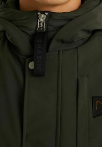 Veste isolante vert foncé avec fermeture éclair, poche à rabat et étiquette en tissu noire. Présente un design cousu et un accent de logo orange.