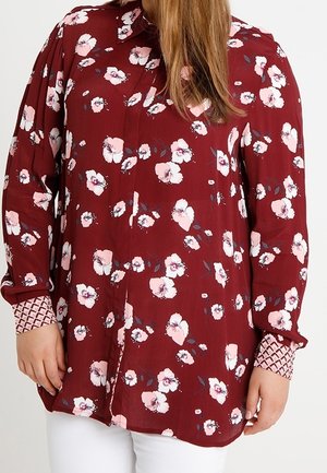 Kobieta nosząca bordową bluzkę z długim rękawem, z różowym i białym kwiatowym nadrukiem oraz wzorzystymi mankietami, w połączeniu z białymi spodniami.