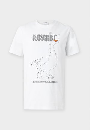 Άνδρας που φοράει μαύρο καπέλο, λευκό t-shirt Moschino με πουά σχέδιο χήνας, μπλε τζιν και μαύρα αθλητικά παπούτσια, στέκεται με το ένα χέρι στην τσέπη.