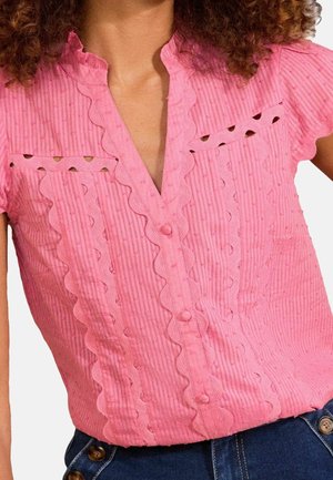 Donna che indossa una blusa rosa testurizzata con bordi smerlati e dettagli con bottoni, abbinata a pantaloni di jeans blu a vita alta con bottoni.