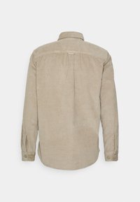 Beige kordfløjls-skjorte med en rygskæring, lange ærmer med knapper og afrundet kant. Karakteristisk ribbet tekstur over det hele.