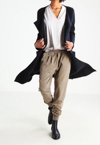 Larga chaqueta negra, camiseta de pico gris claro, pantalones beige y botas de tobillo negras. La chaqueta tiene una textura suave; los pantalones tienen un corte relajado.