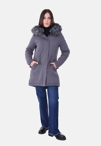 Parka grigia con cappuccio foderato in pelliccia, chiusura con zip, due tasche laterali e vita regolabile con cordino, indossata sopra jeans larghi blu e scarpe nere.