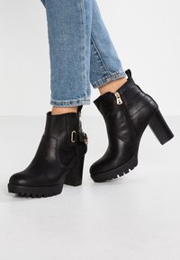 Sorte læder ankelstøvler med kraftige hæle, hurtig lukning i siden og spænde detaljer, iført med opsmøgede blå jeans på en hvid gulv.