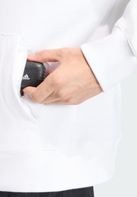 Λευκό φούτερ με φερμουάρ σε τσέπη, στο οποίο φαίνεται ένα χέρι να τραβάει ένα μαύρο αντικείμενο με ανάγλυφο λογότυπο, τονίζοντας το υφή υλικό.