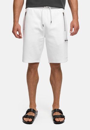 Uomo che indossa pantaloncini bianchi alla lunghezza del ginocchio con tasche con cerniera e sandali neri con fibbia, in piedi davanti a uno sfondo semplice.