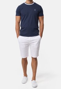 Camiseta de cuello redondo en color navy con ribetes blancos, fabricada en algodón. Combinada con pantalones cortos blancos y zapatos sin cordones, presenta un atuendo casual de verano.