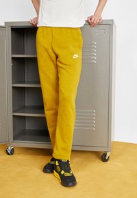 Žluté tepláky s logem Nike, vyrobené z měkké látky. Design zahrnuje elastický pás a zúžené nohavice, doplněné o černé tenisky.