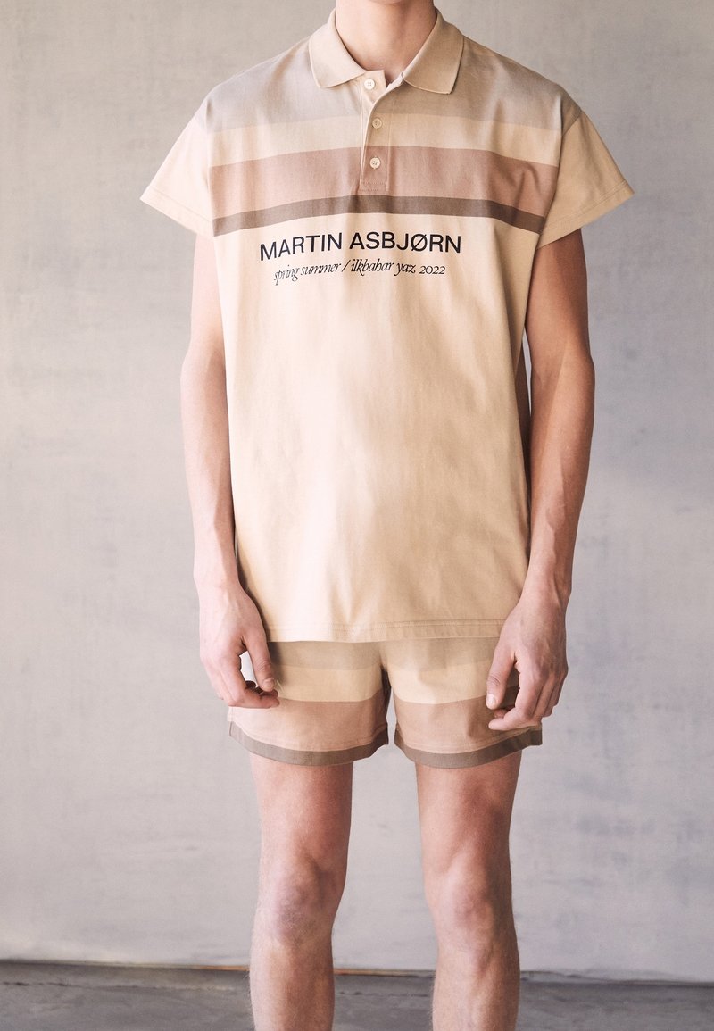 Beige bomullspolo med korta ärmar, med horisontella ränder i dämpade färger och svart textbranding. Matchas med randiga shorts.