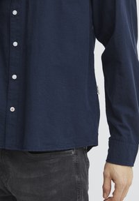 Camicia a maniche lunghe blu navy con tessuto testurizzato, dotata di bottoni bianchi e di un'etichetta laterale; abbinata a jeans grigi scuri.