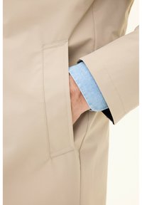 Beige jacket with a sleek design, featuring a side pocket. Cuffs reveal a light blue fabric underneath, showcasing a contrasting texture.