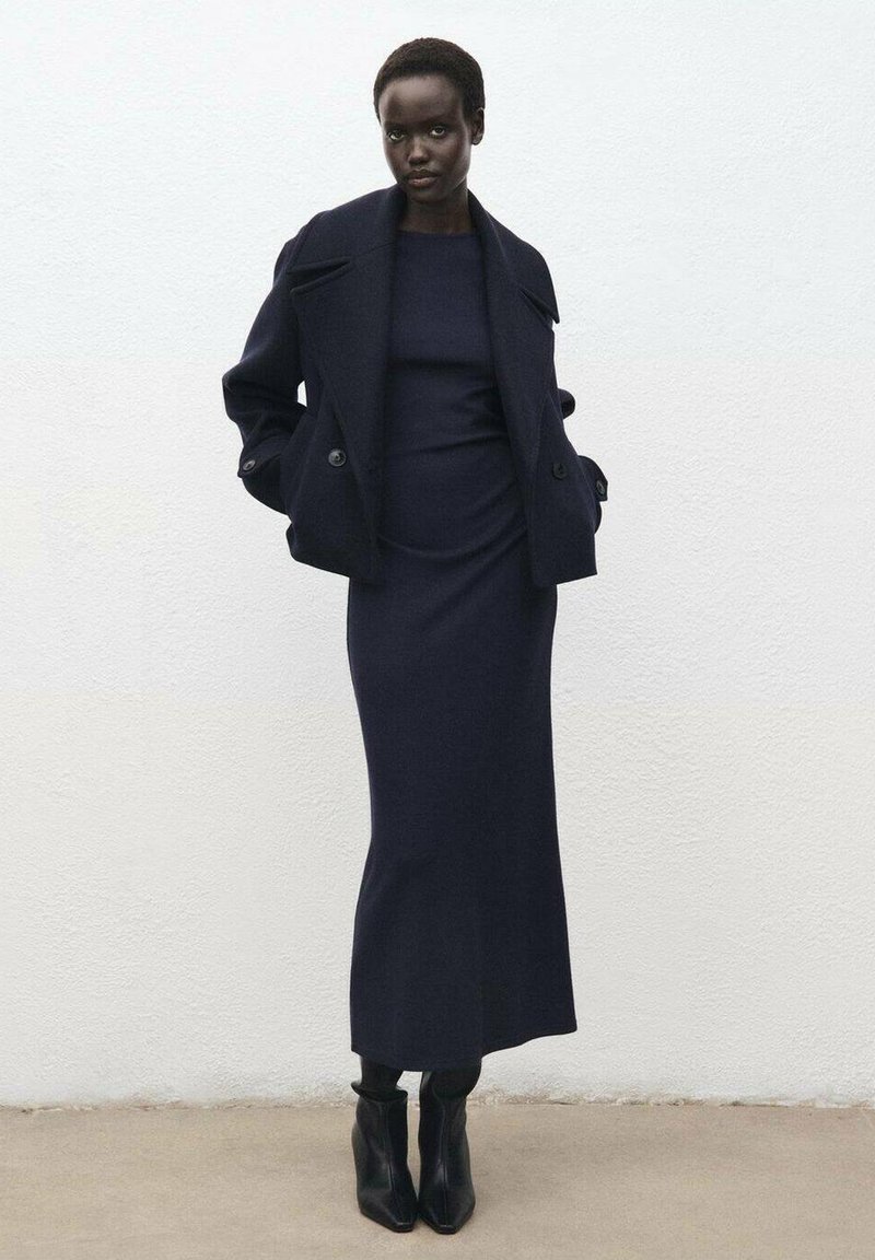 Cappotto in lana blu navy con bottoni doppi, indossato sopra un vestito aderente coordinato, abbinato a stivali neri con punta a gamba. Texture liscia, design minimalista.