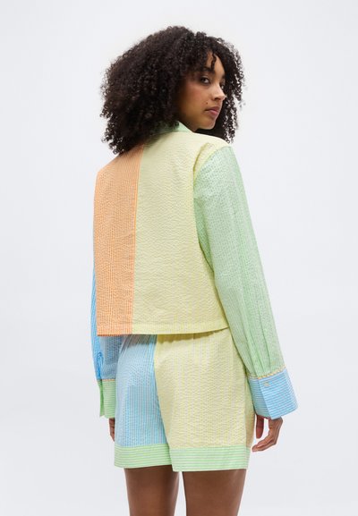 Γυναίκα με σγουρά μαλλιά, που φοράει ένα υφασμάτινο σύνολο σε παστέλ αποχρώσεις με μπλοκ χρωμάτων σε πράσινο, κίτρινο, μπλε και πορτοκαλί, στέκεται μπροστά σε ένα λευκό φόντο.