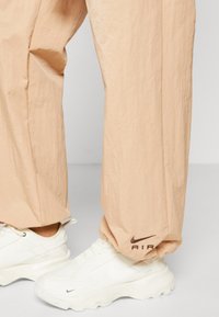 Kalhoty z lehké, krčící se látky v broskvové barvě s tmavě hnědým logem "AIR" na spodním lemu, doplněné bílými botami Nike.