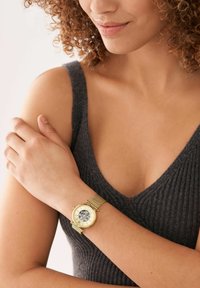 Eine goldene Armbanduhr mit einem Mesh-Armband, runder Gehäuseform und einem klaren Ziffernblatt mit römischen Ziffern, getragen an einem nackten Arm, vor einem grauen Oberteil.