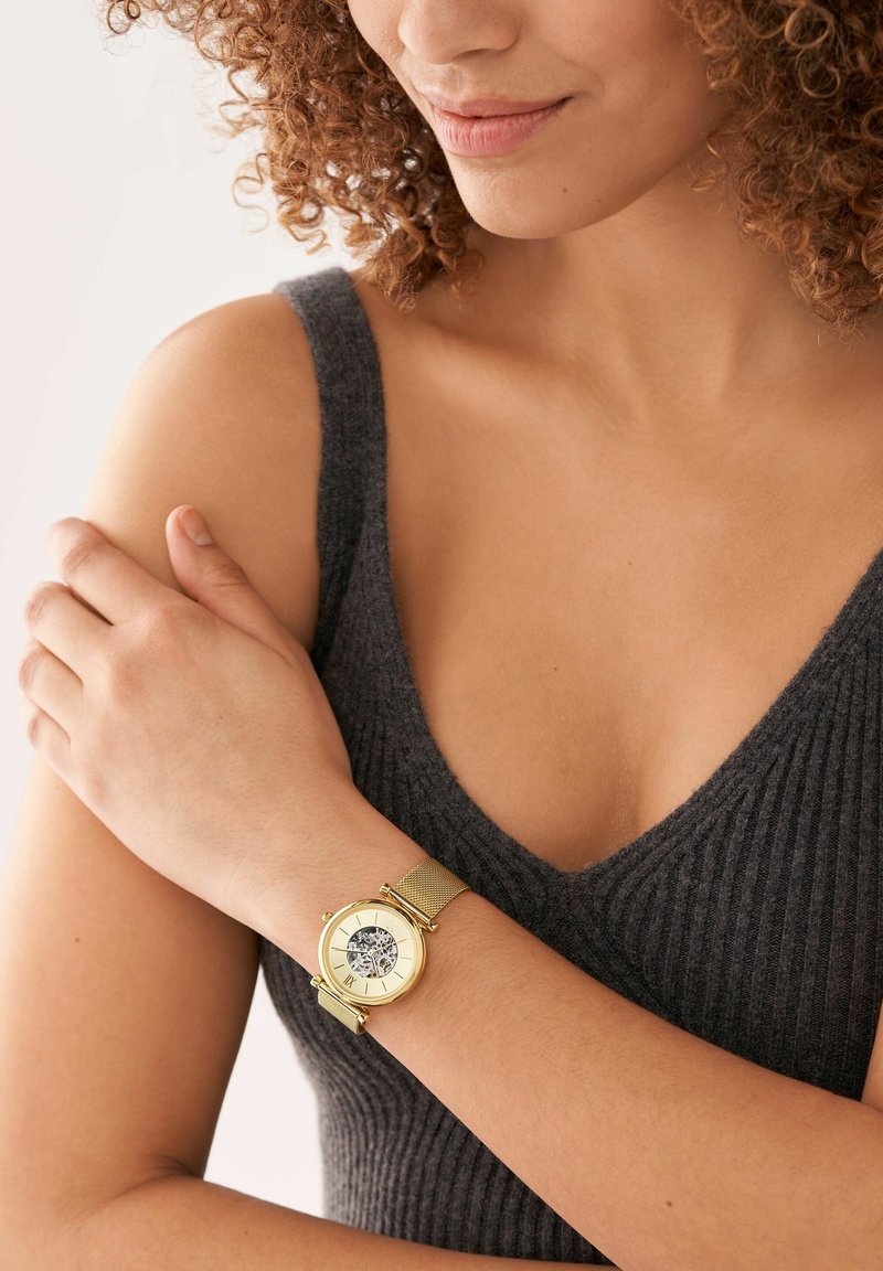 Eine goldene Armbanduhr mit einem Mesh-Armband, runder Gehäuseform und einem klaren Ziffernblatt mit römischen Ziffern, getragen an einem nackten Arm, vor einem grauen Oberteil.