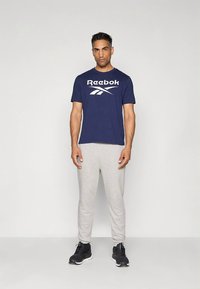 Marineblå Reebok t-shirt med hvidt logo, kombineret med lysegrå sweatpants. Fodtøjet er sorte sneakers. Enkel, afslappet sportstøj.