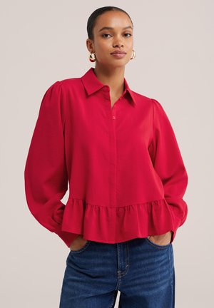 Femme portant une blouse rouge à manches longues avec un col et un ourlet volanté, associée à un jean bleu, debout les mains dans les poches.