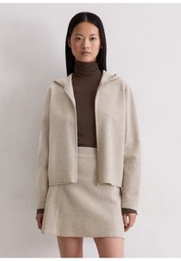 Beige zip-up jack met een capuchon en een zachte textuur, gecombineerd met een bruine coltrui en bijpassende rok, wat resulteert in een samenhangende outfit.