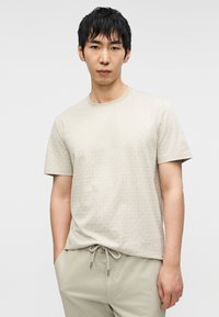 Μπεζ T-shirt με κοντά μανίκια και διακριτικό σχέδιο, στρογγυλή λαιμόκοψη και απαλή υφή. Συνδυάζεται με ανοιχτό μπεζ παντελόνι με κορδόνι στη μέση.