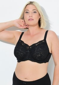 Černá krajková podprsenka s nastavitelnými ramínky, květinovým vzorem, s výztuží a vlnitými okraji. Přímo vepředu se nachází malá mašlička uprostřed.