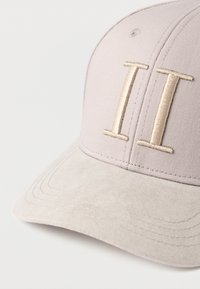 Világos mályvaszínű baseball sapka bézs színű velúr napellenzővel. Az elején arany "II" hímzés, és varrott díszítőelemek a perem mentén.