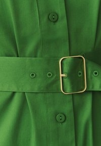 Zelený textilný opasok so zlatou obdĺžnikovou prackou pripnutý cez zodpovedajúci zelený odev s viditeľnými gombíkmi a detailmi švov.