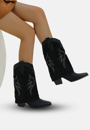 Botas de cowboy de ante negro con punteras puntiagudas y tacones cortos en bloque. Presentan un bordado decorativo plateado y lengüetas en el eje.
